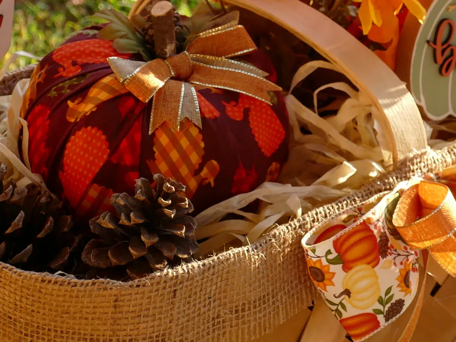 Close view of a deep red patterned fabric pumpkin with a gold ribbon bow and natural wood stem, resting among pinecones and burlap.