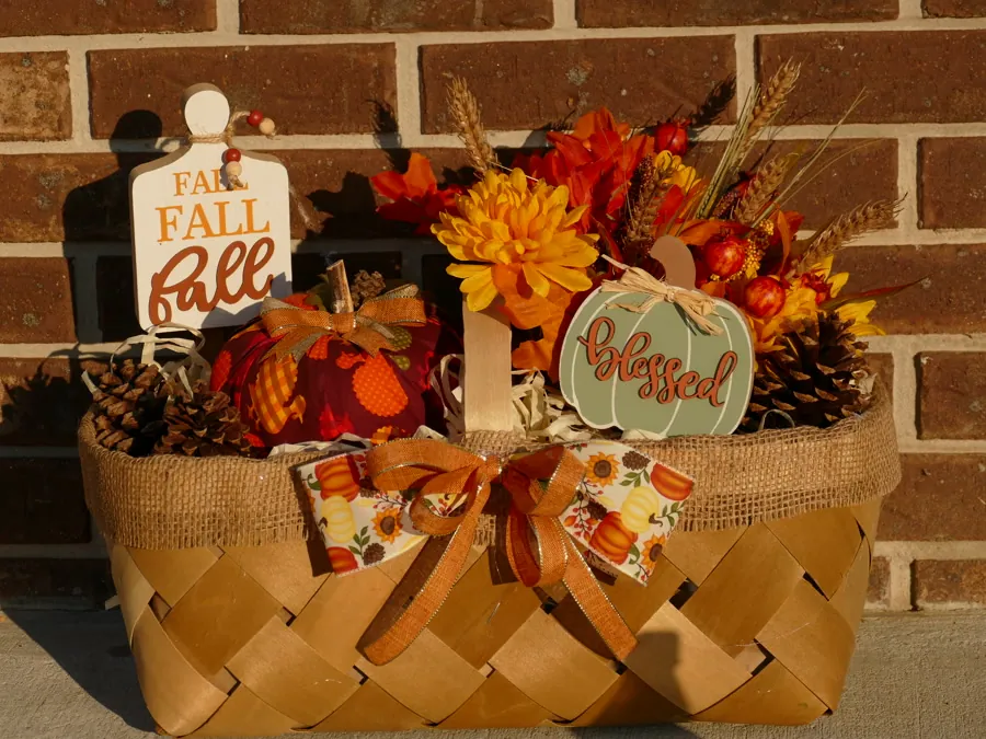 Woven basket wrapped in burlap holding a red fabric pumpkin, pinecones, orange and gold mums, wheat stems, and a sage green pumpkin sign reading 'blessed'.