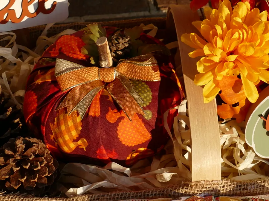 Deep red patterned fabric pumpkin with a gold bow and wood stem, surrounded by pinecones and paper filler inside a woven basket.